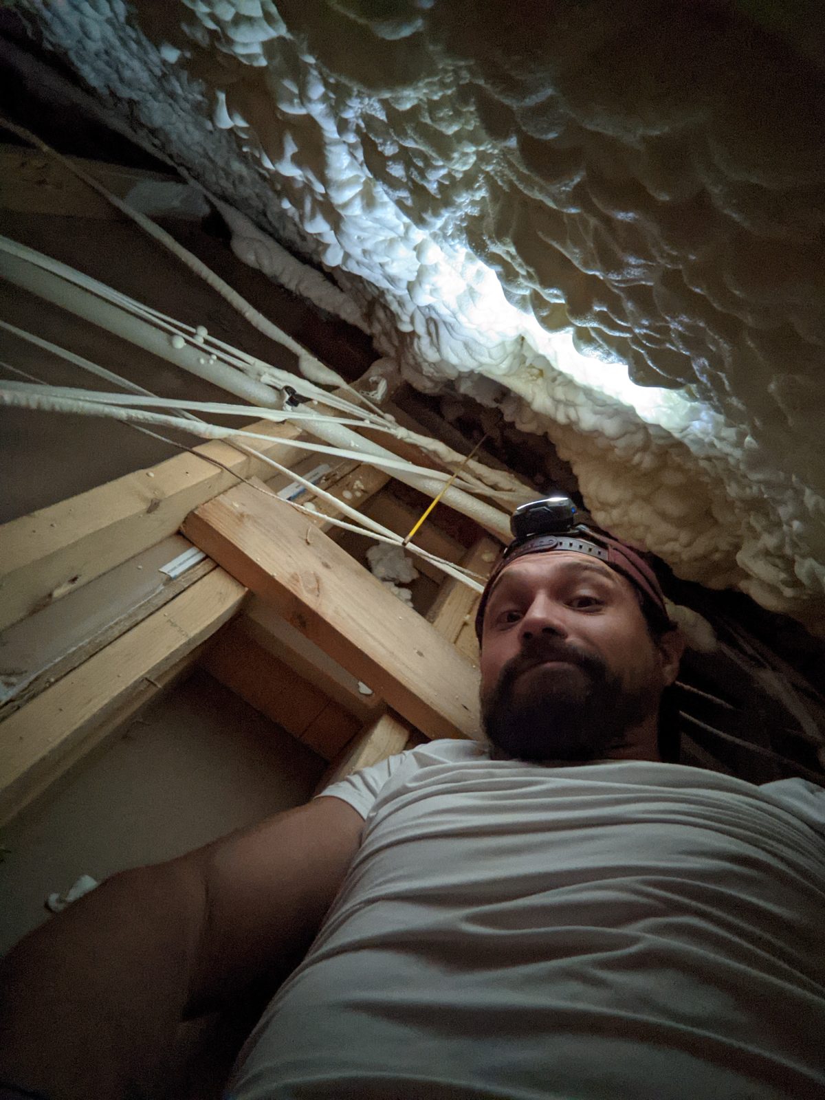Steven wearing a headlamp, lying under spray foam insulation while running wire through an attic