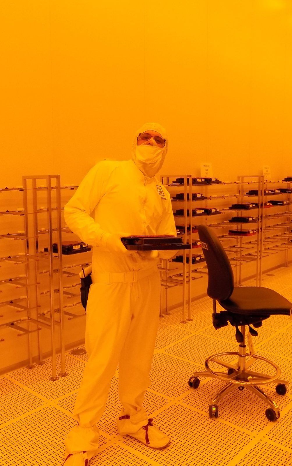 Steven in a full cleanroom suit holding a wafer carrier in Intel's yellow-lit fab